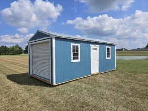 A Slate Blue Premier Portable Buildings Garage with white trim and a roll-up garage door open, revealing a wooden interior, set in a green grassy field under a blue sky with white clouds.