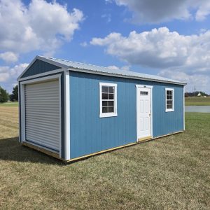 A Slate Blue Premier Portable Buildings Garage with white trim and a roll-up garage door open, revealing a wooden interior, set in a green grassy field under a blue sky with white clouds.