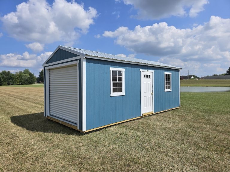 A Slate Blue Premier Portable Buildings Garage with white trim and a roll-up garage door open, revealing a wooden interior, set in a green grassy field under a blue sky with white clouds.