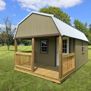 A cozy olive-colored Premier Lofted Barn Cabin with a white metal roof and a wooden front porch, situated in a grassy field surrounded by green trees under a clear blue sky.