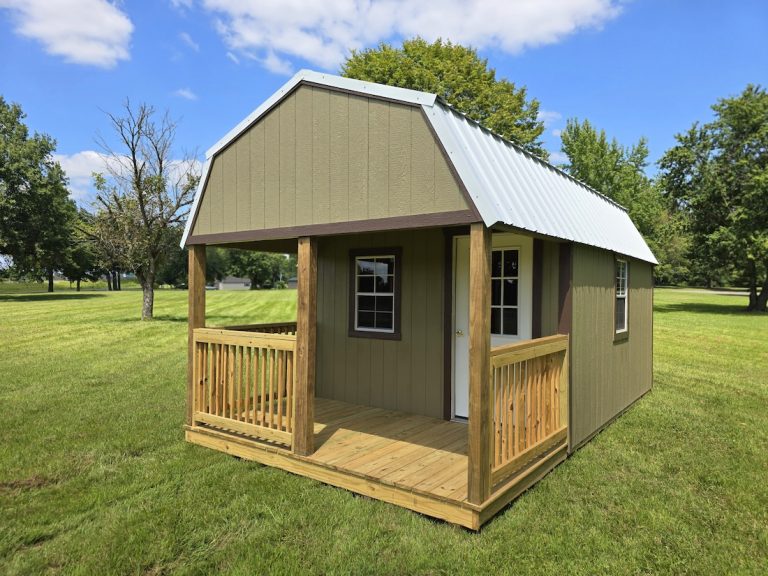A cozy olive-colored Premier Lofted Barn Cabin with a white metal roof and a wooden front porch, situated in a grassy field surrounded by green trees under a clear blue sky.