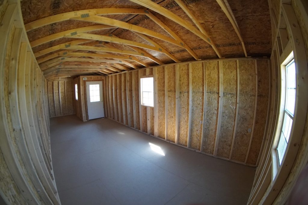 Interior view of a Premier Side Cabin featuring exposed timber framing, oriented strand board (OSB) sheathing on walls and ceiling, a white paneled door at the far end, and two windows providing natural light.