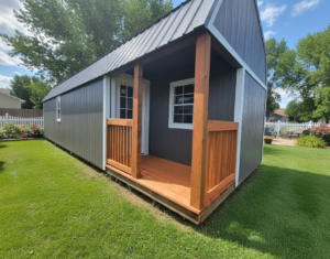 Image of a Gray with White Trim Premier Side Lofted Barn Cabin in a back yard; surrounded by lush green grass, a blue sky with wispy clouds, and a white picket fence around the edge of the yard.