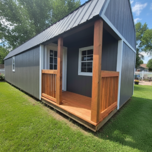 Image of a Gray with White Trim Premier Side Lofted Barn Cabin in a back yard; surrounded by lush green grass, a blue sky with wispy clouds, and a white picket fence around the edge of the yard.
